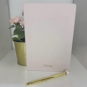 Könnte beinhalten: Ein hellrosa Notizbuch mit dem Namen "Elena" in goldfarbenen Buchstaben. Ein goldener Stift mit Perlenspitze liegt vor dem Notizbuch. Ein kleiner goldener Topf mit rosa Blumen befindet sich links vom Notizbuch.
