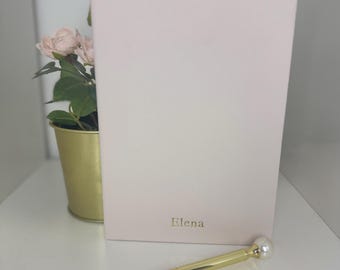 Puede incluir: Un cuaderno rosa claro con el nombre "Elena" en letras doradas. Un bolígrafo dorado con punta de perla descansa frente al cuaderno. Una pequeña maceta dorada con flores rosas está a la izquierda del cuaderno.