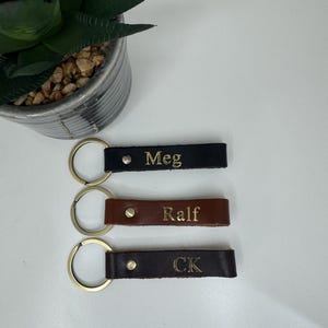 May include: Three leather keyrings with gold key rings. The keychains are black, brown, and dark brown. The names "Meg", "Ralf", and "CK" are embossed in gold on the leather.