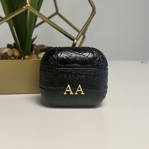 May include: Black textured case with gold-colored initials "AA". The case is likely for earbuds or a similar small electronic device. The case has a rounded shape and a textured surface. The initials are centered on the front of the case.