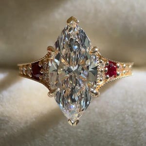 May include: A gold ring featuring a large marquise-cut center stone, flanked by two small round red stones on each side. The band is detailed with small accent stones and a textured, milgrain-style edge along the setting of the center stone.