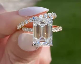 May include: A collection of three gold rings. The primary ring features a large, rectangular, clear gemstone. Two additional rings are visible, one with small, round gemstones and another with a band of smaller stones. The rings are held against a blurred green background.