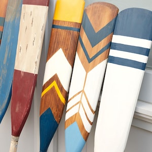 Può includere: Una collezione di remi decorativi in legno in vari colori e motivi. I remi presentano disegni in blu, bianco, rosso, giallo e marrone. Alcuni hanno motivi a chevron e a strisce, altri blocchi di colore pieno. Adatti per decorazioni nautiche o costiere.