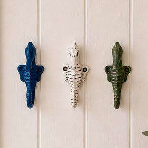 Puede incluir: Tres ganchos de pared con forma de caballito de mar montados verticalmente en fila. Los ganchos son de color azul oscuro, blanco y verde oscuro, cada uno con dos orificios para tornillos. El diseño incluye detalles texturizados.