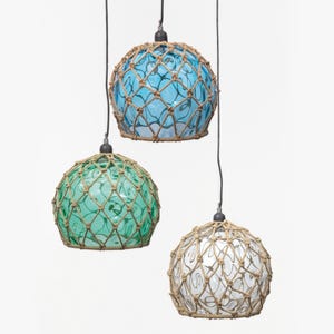 Op de afbeelding: Drie hangende pendelverlichting met bolvormige glazen kappen in blauw, groen en wit. Elke kap is omhuld met een geweven touwnet. De lampen hangen aan zwarte snoeren tegen een witte achtergrond, wat een nautisch thema creëert.