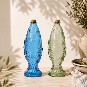 May include: Two glass bottles shaped like fish, one in vibrant blue and the other in light green. Each bottle features a textured scale pattern, a circular eye detail, and a flared tail base. Both bottles are topped with a small, cylindrical cork stopper.
