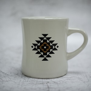 May include: White ceramic mug with a black and brown geometric design. The design features a diamond shape with a cross in the centre.
