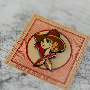 May include: An enamel pin featuring a woman wearing a brown cowboy hat and a red bandana. The pin is on a card with the text "Rock & Rose Motel".