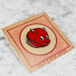 May include: Red enamel pin featuring a cartoon devil with horns and a smile. The pin is on a card with the text "SICK & ROSE MOTEL".