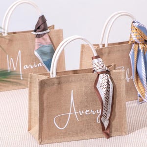 May include: Three jute tote bags with white handles, each bearing a name in cursive script. The bags are accessorized with patterned scarves in different colours, adding a decorative element.