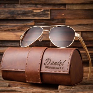 May include: Gold aviator sunglasses with brown lenses rest on a brown leather case. The case is embossed with the name "Daniel" and the word "Groomsman". The sunglasses have gold frames and wooden arms.