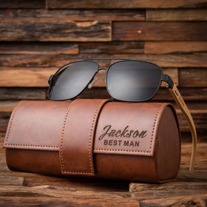 May include: Black aviator sunglasses with a brown leather case. The case is embossed with the name "Jackson" and the words "BEST MAN". The sunglasses have a black frame and dark lenses. The case is approximately 7 inches long.