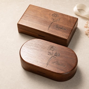 May include: Two dark brown wooden boxes with hinged lids. One box is rectangular and engraved with a flower and the name Sophia. The other box is oval and engraved with a flower and the name Layla. Both feature smooth, polished wood grain surfaces.