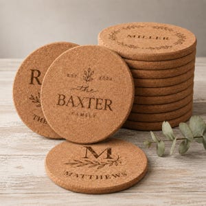 May include: A stack of round cork coasters with engraved text and botanical designs. One coaster features the text "EST. 2024 the BAXTER FAMILY" with a leaf sprig. Another shows "M MATTHEWS" with a leafy branch. The coasters have a natural brown texture.