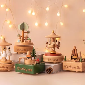 May include: A collection of wooden music boxes with personalized names, including a carousel, a tree with rabbits, and a train scene. The boxes are in various shapes and sizes, with a Christmas tree and lights in the background.