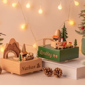 May include: Two wooden music boxes with Christmas themes. One is light brown with "Nathan" engraved, featuring a train and mountain design. The other is green with "Lindley" engraved, showcasing a house and tree. Christmas trees and pinecones are in the background.