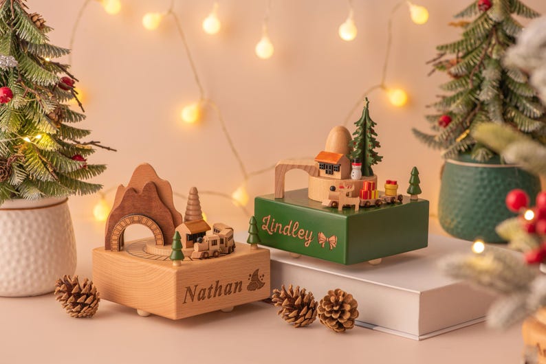 May include: Two wooden music boxes with Christmas themes. One is light brown with "Nathan" engraved, featuring a train and mountain design. The other is green with "Lindley" engraved, showcasing a house and tree. Christmas trees and pinecones are in the background.
