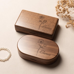 Può includere: Due scatole in legno con finiture lisce. La scatola rettangolare presenta una rosa incisa e il nome Luna. La scatola ovale presenta un fiore inciso e il nome Elena. Entrambe le scatole hanno una venatura del legno marrone scuro e un coperchio.