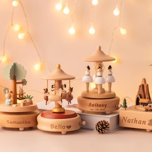 May include: Wooden music boxes with personalized names, including a carousel, a tree with rabbits, and a ballerina design. The boxes are light brown with decorative elements and are set against a warm, festive background.