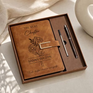 May include: A tan notebook with a gold-toned metal clasp, featuring the text THE NEXT Chapter, SARAH WILSON, and Each chapter ushers in new growth, alongside a floral illustration. The set includes a matching pen and a refill in a brown box.