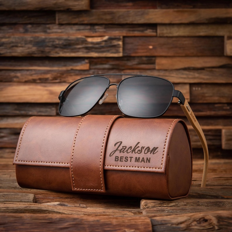 May include: Black aviator sunglasses with a brown leather case. The case is embossed with the name "Jackson" and the words "BEST MAN". The sunglasses have a black frame and dark lenses. The case is approximately 7 inches long.