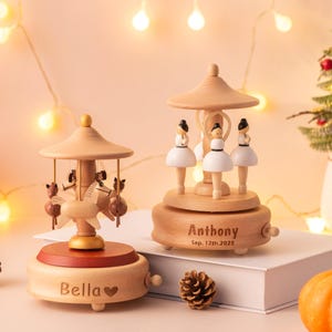 May include: Two wooden music boxes with carousel designs. One features ballerina figures, the other has horse figures. The music boxes are personalized with names and dates. A small Christmas tree and pumpkins are also in the image.