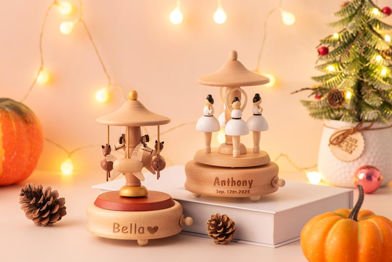 May include: Two wooden music boxes with carousel designs. One features ballerina figures, the other has horse figures. The music boxes are personalized with names and dates. A small Christmas tree and pumpkins are also in the image.