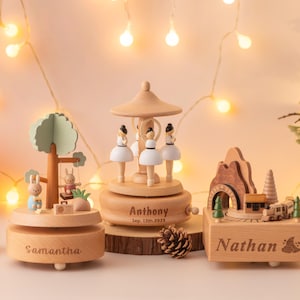 May include: Wooden music boxes with personalized names and dates. One features a carousel with ballerina figures, another a tree with rabbits, and the third a train scene. A small Christmas tree and pumpkin are also in the image.
