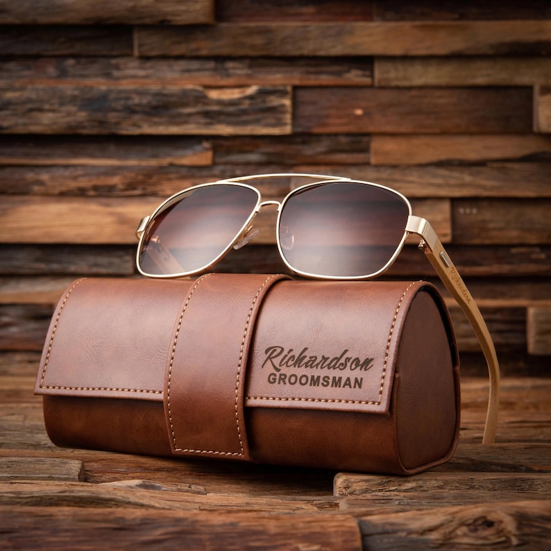 May include: Gold-framed sunglasses with brown lenses rest on a brown leather case. The case has a strap and the text "Richardson GROOMSMAN" is visible. The sunglasses have a classic aviator design.
