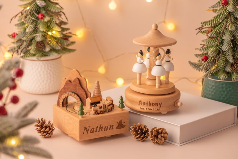 May include: Wooden music boxes with personalized names and dates. One features a train scene, the other a carousel with ballerinas. Christmas trees and pinecones decorate the scene. The carousel is engraved with "Anthony Sep. 12th, 2025".