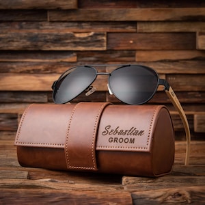 May include: Black aviator sunglasses with a brown leather case. The case is embossed with the name "Sebastian" and the word "Groom". The sunglasses have a black metal frame and dark lenses. The case is a cylindrical shape.
