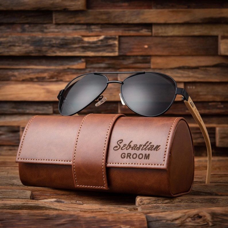 May include: Black aviator sunglasses with a brown leather case. The case is embossed with the name "Sebastian" and the word "Groom". The sunglasses have a black metal frame and dark lenses. The case is a cylindrical shape.