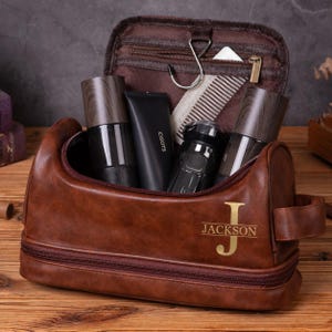 May include: A brown leather wash bag with a gold monogram featuring the letter J and the name JACKSON. The open bag contains two dark wood-patterned bottles, a black tube labelled CISOTS, a black electric shaver, and a metal hanging hook.