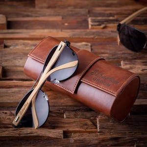 May include: Sunglasses with a brown leather case. The sunglasses have black frames and light-colored wooden arms. The case is embossed with the name "Sebastian" and the word "Groom". The background is a wooden surface.