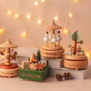 May include: A collection of wooden music boxes with personalized names, including a carousel, ballerinas, a tree with rabbits, and a train scene. The music boxes are light brown with red and green accents, and are displayed with Christmas trees and fairy lights.