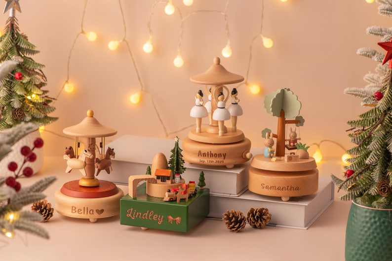 May include: A collection of wooden music boxes with personalized names, including a carousel, ballerinas, a tree with rabbits, and a train scene. The music boxes are light brown with red and green accents, and are displayed with Christmas trees and fairy lights.