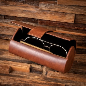 May include: A brown leather eyeglasses case with a black interior, open to reveal a pair of gold-rimmed sunglasses. The case is cylindrical with a flap closure. The background is a wooden surface.