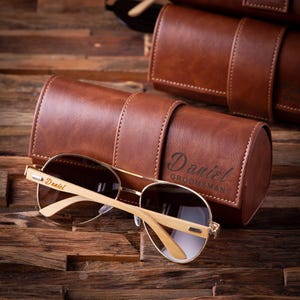May include: Sunglasses with gold frames and light wood arms rest near a brown leather case. The case is embossed with the name "Daniel" and the word "Groomsman". The sunglasses also have the name "Daniel" on the arm.