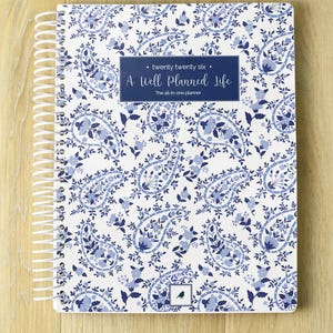 May include: A spiral-bound planner with a white and blue paisley pattern. The cover features the text "twenty twenty six" and "A Well Planned Life". The planner is on a light wood surface.