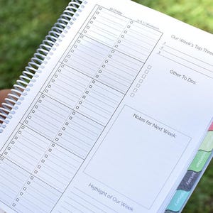 Puede incluir: Una agenda en espiral blanca con p&aacute;ginas rayadas. Incluye secciones para tareas diarias, objetivos semanales y notas. Las pesta&ntilde;as laterales est&aacute;n etiquetadas con los meses. La p&aacute;gina abierta muestra el texto "Our Week's Top Three".