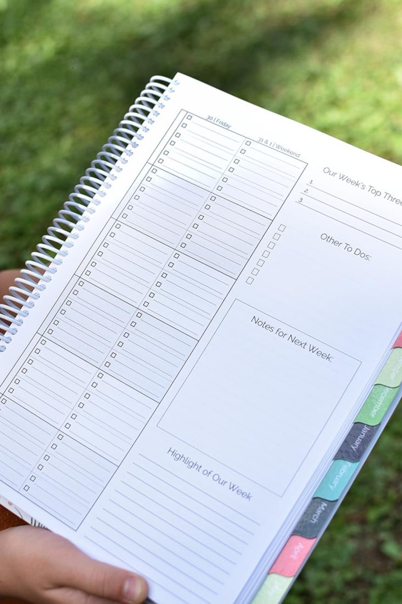 Puede incluir: Una agenda en espiral blanca con p&aacute;ginas rayadas. Incluye secciones para tareas diarias, objetivos semanales y notas. Las pesta&ntilde;as laterales est&aacute;n etiquetadas con los meses. La p&aacute;gina abierta muestra el texto "Our Week's Top Three".
