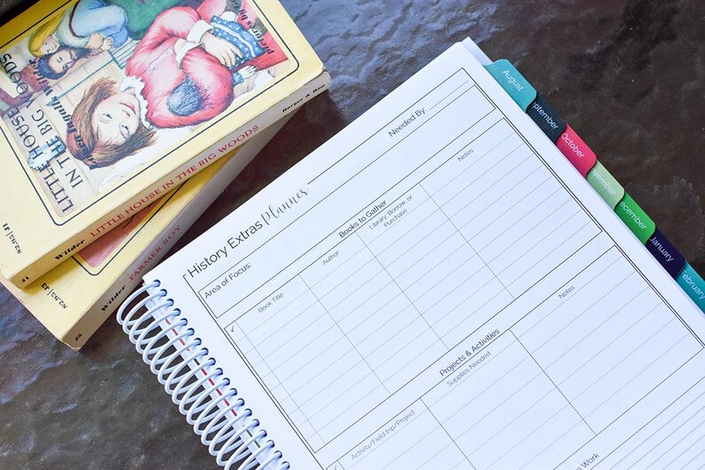 Puede incluir: Un planificador encuadernado en espiral con el t&iacute;tulo "History Extras Planner" y una pila de libros antiguos. El planificador tiene separadores con pesta&ntilde;as y p&aacute;ginas rayadas para notas. Los libros se titulan "Little House in the Big Woods" y "Farmer Boy".