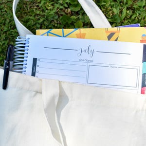 Puede incluir: Un bolso tote de lona blanca con una agenda con espiral y un bol&iacute;grafo negro. La agenda est&aacute; abierta en una p&aacute;gina con la etiqueta "julio" con espacio para objetivos y un tema. Otros cuadernos son visibles dentro de la bolsa.