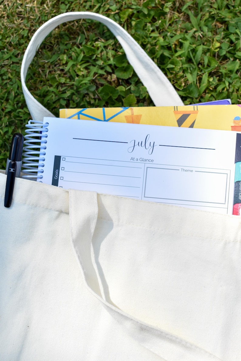 Puede incluir: Un bolso tote de lona blanca con una agenda con espiral y un bol&iacute;grafo negro. La agenda est&aacute; abierta en una p&aacute;gina con la etiqueta "julio" con espacio para objetivos y un tema. Otros cuadernos son visibles dentro de la bolsa.