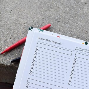 Puede incluir: Un cuaderno "School Year Prep List" con un borde floral, abierto en una p&aacute;gina de lista de verificaci&oacute;n. Un bol&iacute;grafo rojo y un bol&iacute;grafo negro descansan sobre el cuaderno. El cuaderno incluye el texto "School Year Prep List".