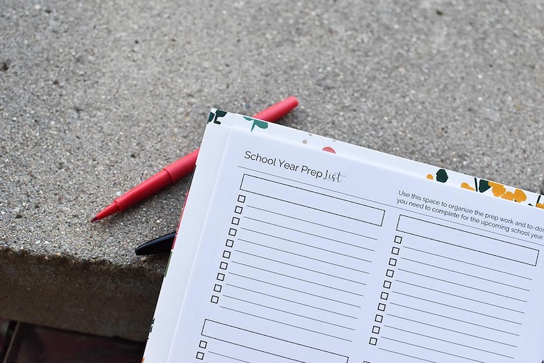 Puede incluir: Un cuaderno "School Year Prep List" con un borde floral, abierto en una p&aacute;gina de lista de verificaci&oacute;n. Un bol&iacute;grafo rojo y un bol&iacute;grafo negro descansan sobre el cuaderno. El cuaderno incluye el texto "School Year Prep List".