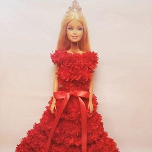 Peut inclure: Une robe rouge à volants, en fourrure, avec un ruban de satin rouge et une couronne sur une poupée.