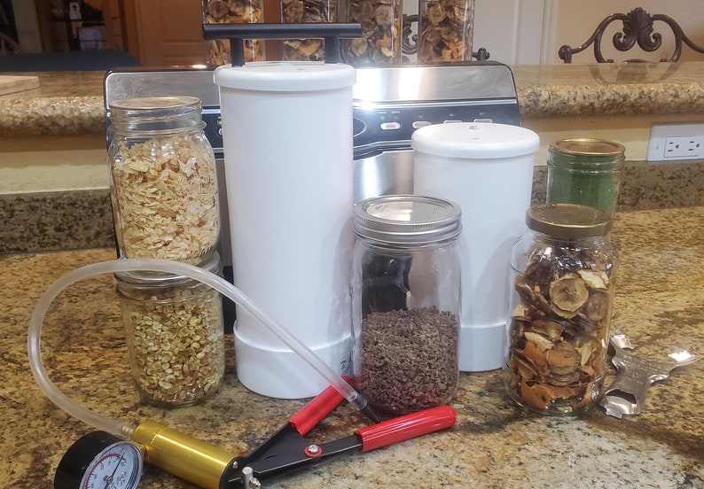 May include: A set of six glass jars with lids, filled with dried fruit and other ingredients. The jars are arranged on a countertop next to a white vacuum sealer and a hand pump.
