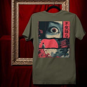 May include: Olive green t-shirt featuring a graphic design with a large eye, a red sun, and Japanese text. The artwork includes a figure and traditional Japanese architecture. The design is in shades of blue, red, and black.