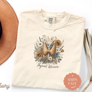 May include: An ivory-colored t-shirt featuring a central graphic of a human thyroid gland surrounded by a detailed arrangement of wildflowers, poppies, and foliage. Below the illustration, the text reads Thyroid Warrior in a cursive script.